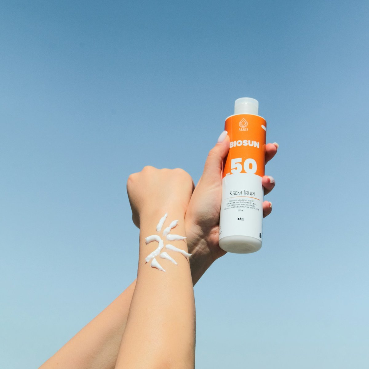 Hands holding an orange-and-white sunscreen bottle against a blue sky, with sunscreen drawn in a sun shape on a forearm.